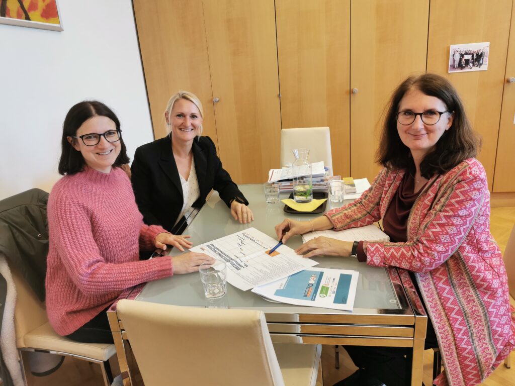 3 Personen (3 Frauen) sitzen an einem Tisch in einem Büroraum und lächeln. Auf dem Tisch liegen Arbeitsblätter und Wassergläser