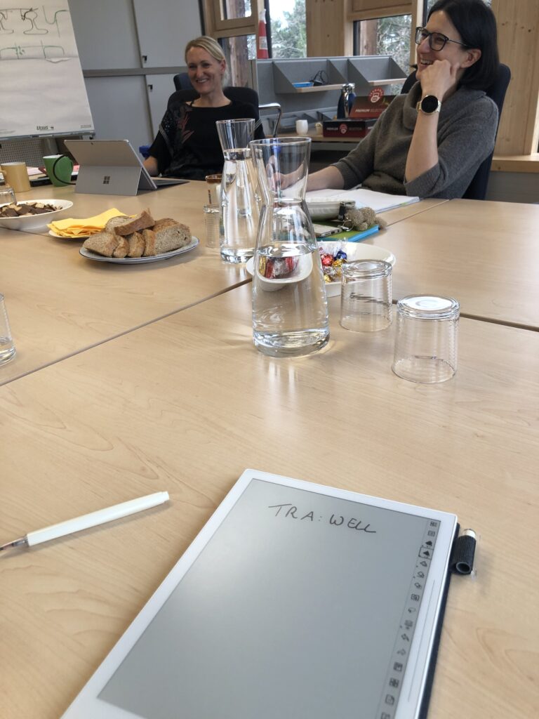meeting room: 2 women laughing, sitting around a table, tablet in front of the picture showing "tra:well" 