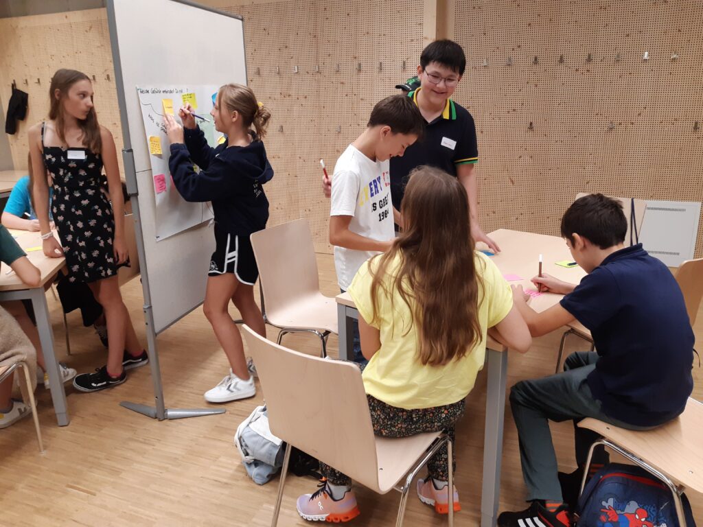 Children sit and stand at the table, a pin board is in the background and is labeled by a pupil.